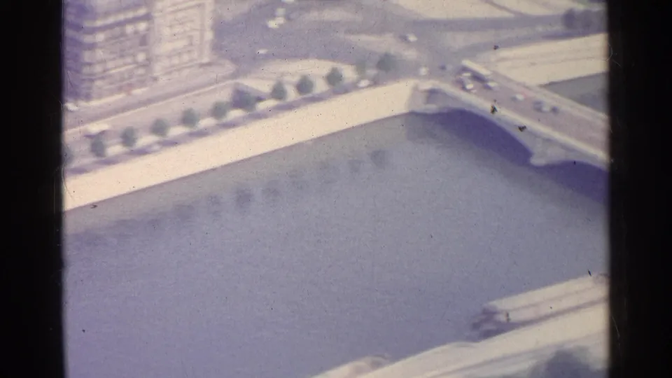 1961 Paris aerial frame showing a bridge over the river, cars crossing above, and riverside buildings and walkways nearby.