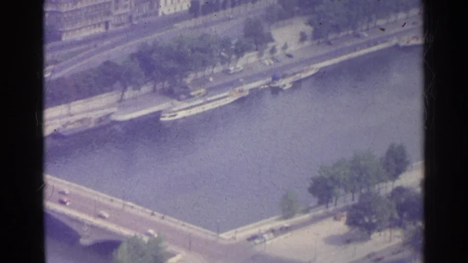 1961 Paris aerial frame with a Seine bridge, riverside boats, tree-lined embankment, and pale buildings in the background.