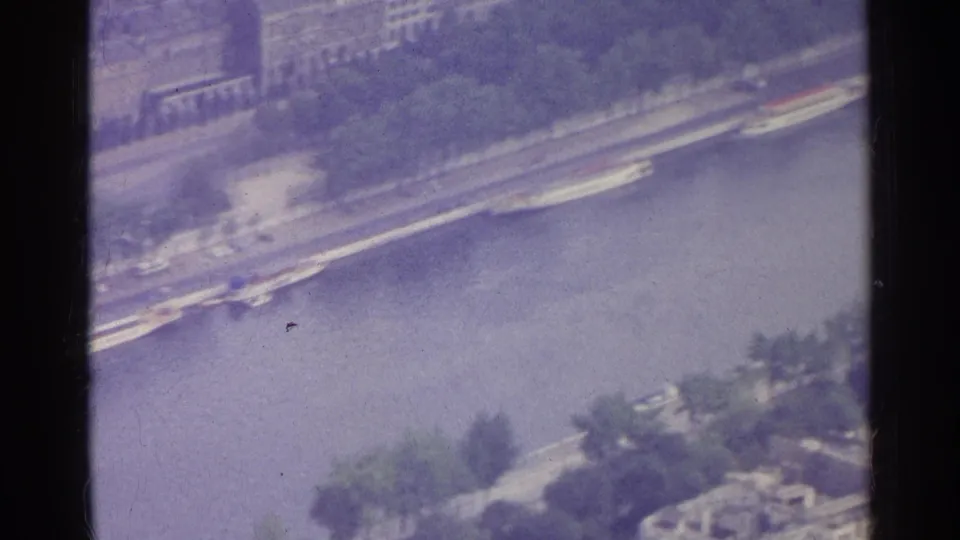 1961 Paris 8mm frame showing long boats along the Seine, a tree-lined riverbank, and urban buildings beyond.