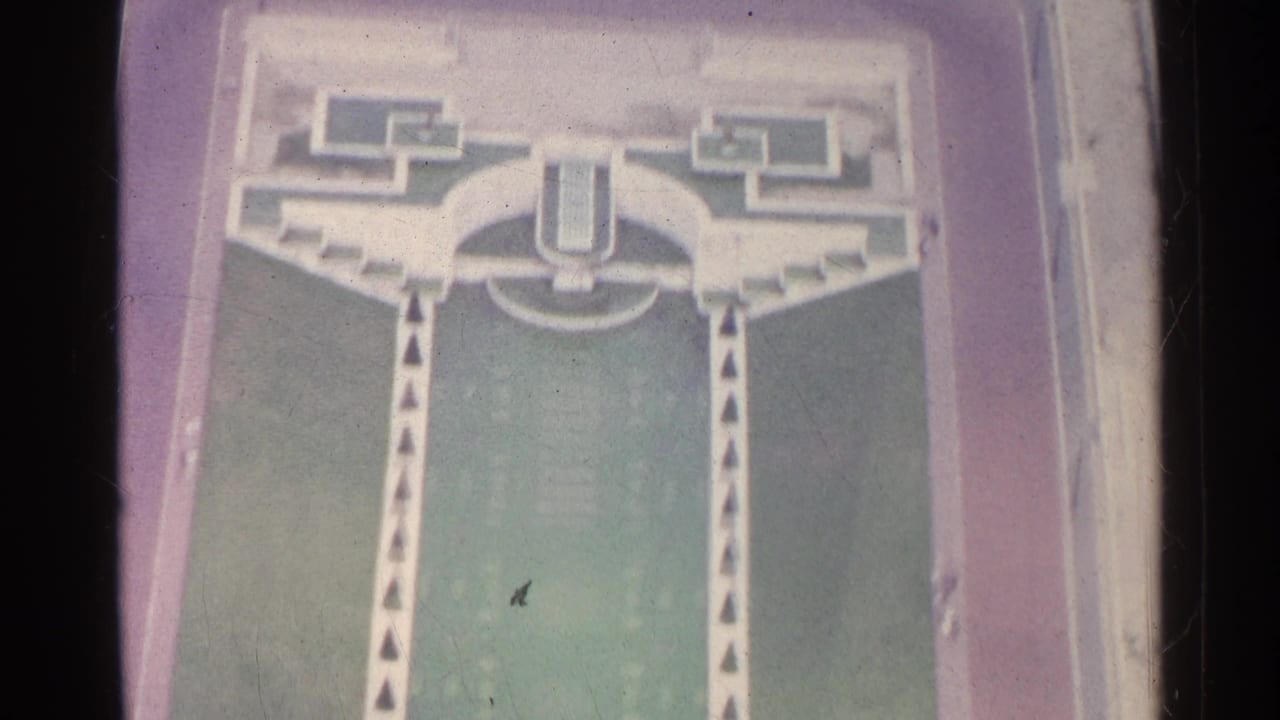 High-angle view of a formal lawn, curved paths, stairs, and monumental building edges.