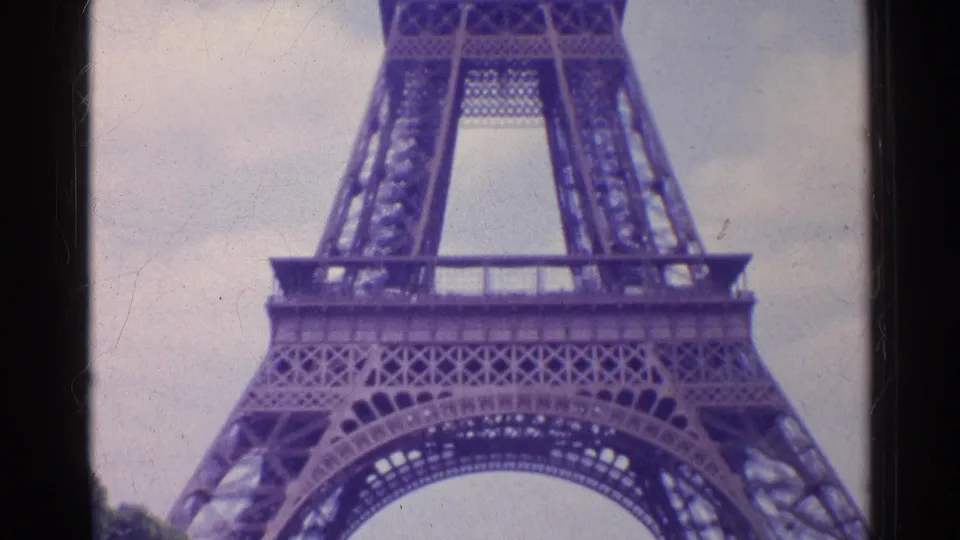1961 Paris 8mm frame showing the Eiffel Tower base, lower arch, and lattice supports beneath a cloudy sky.
