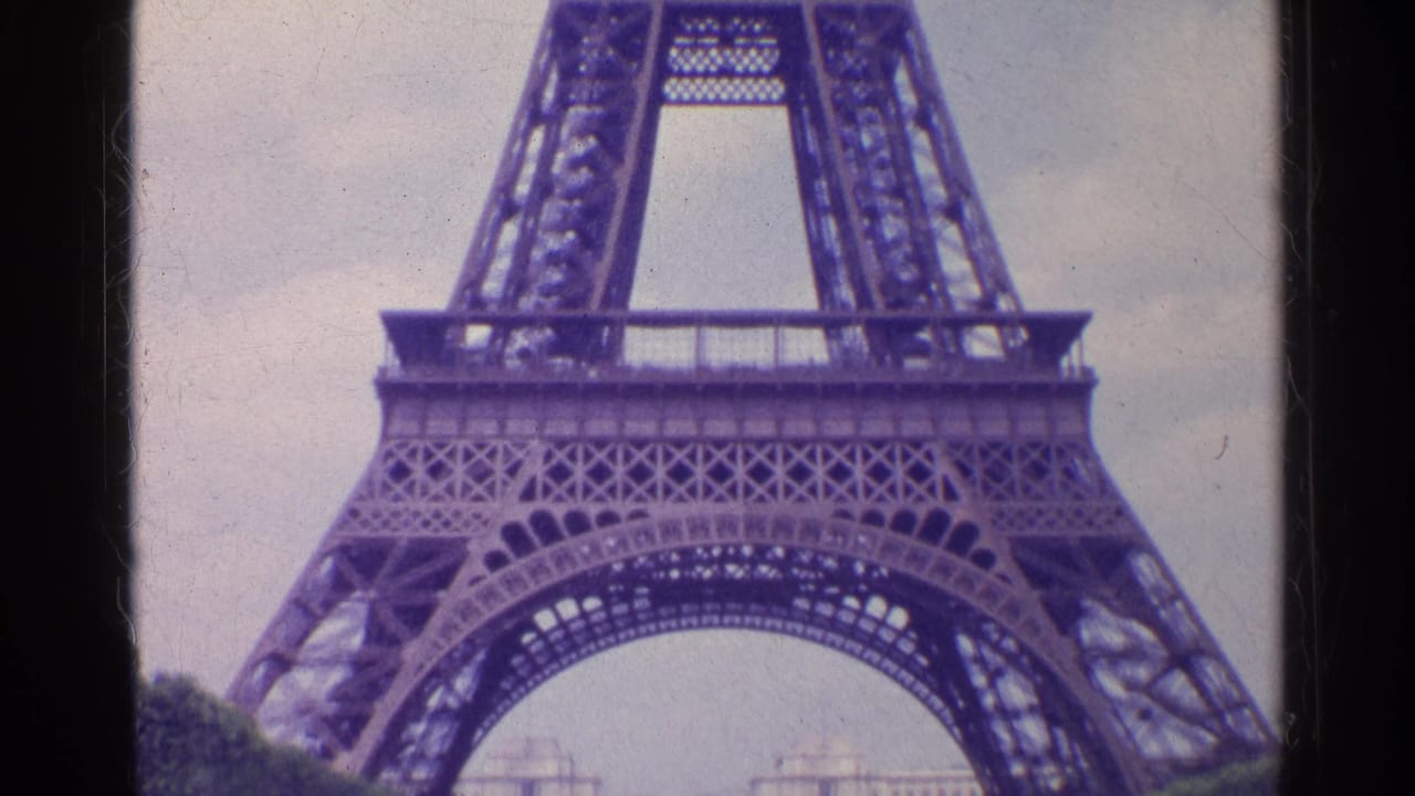1961 Paris home-movie frame showing the Eiffel Tower base and iron lattice from a low angle under a pale sky.
