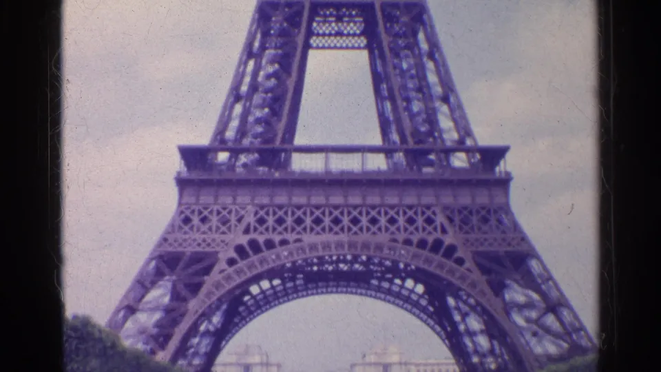 1961 Paris home-movie frame showing the Eiffel Tower base and iron lattice from a low angle under a pale sky.