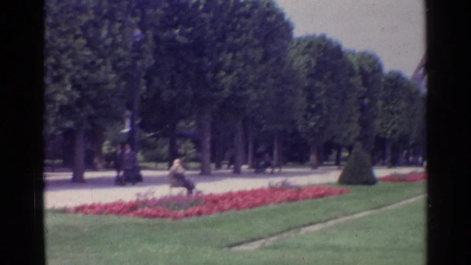 1961 Paris home-movie frame showing a tree-lined park walkway near the Eiffel Tower grounds with flowers, lawn, and visitors.