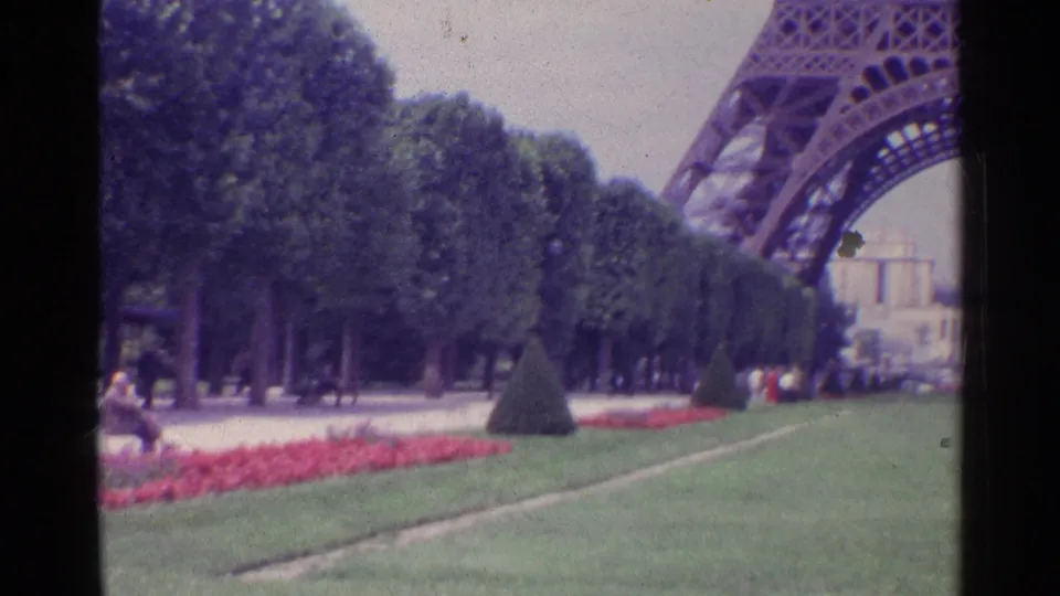 1961 Paris home-movie frame showing lawns, red flower beds, clipped trees, and part of the Eiffel Tower above the grounds.