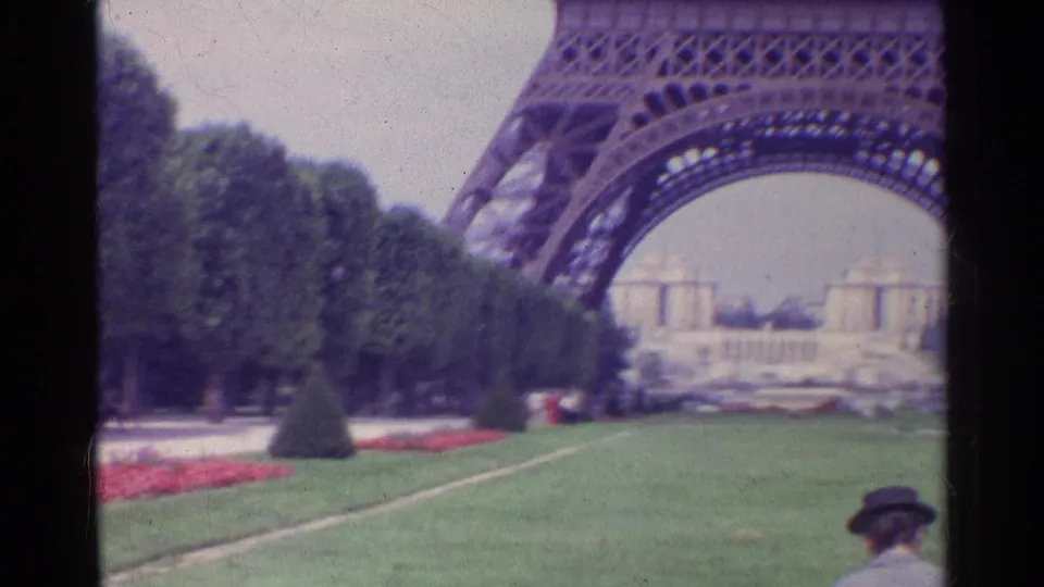 1961 Paris home-movie frame showing the Eiffel Tower arch above formal lawns, red flower beds, distant buildings, and visitors.