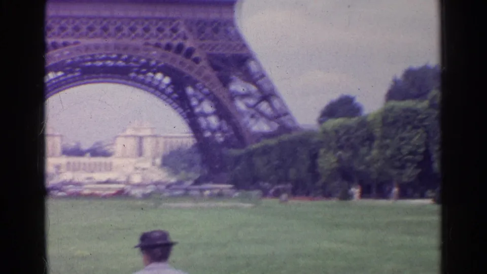 1961 Paris 8mm frame of the Eiffel Tower base from the park, with clipped trees, distant buildings, cars, and a partial visitor figure.