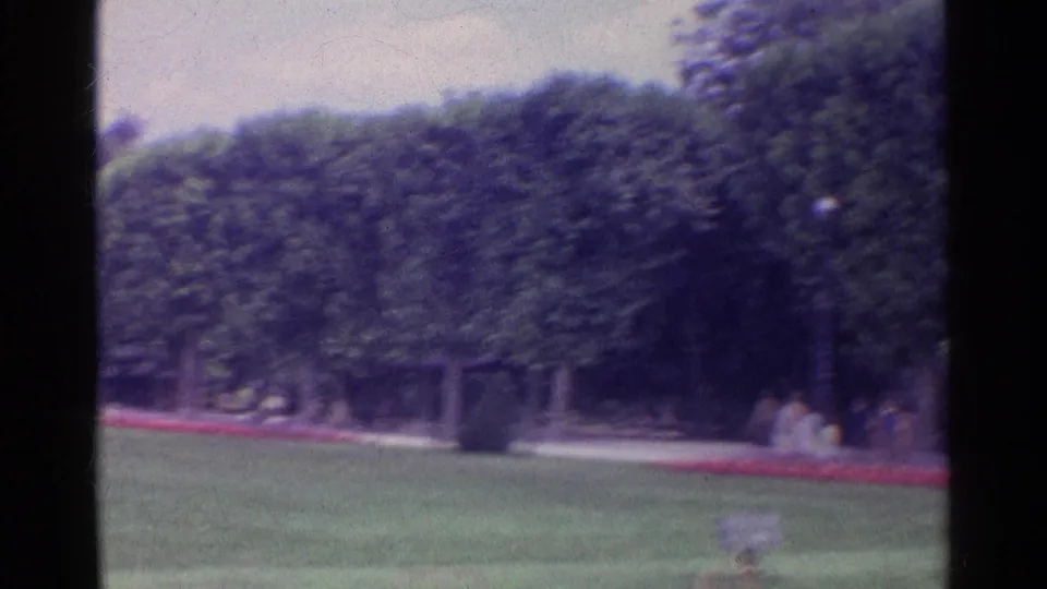 1961 Paris 8mm frame of the formal park near the Eiffel Tower, showing clipped trees, lawn, flower beds, and distant seated visitors.