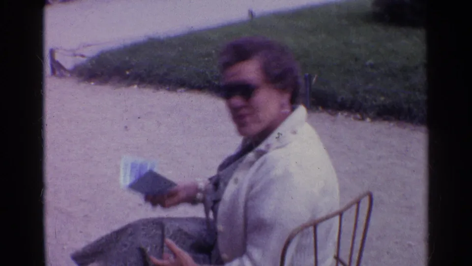 Woman in sunglasses and a light jacket sits on a metal garden chair in a Paris park, holding a booklet.