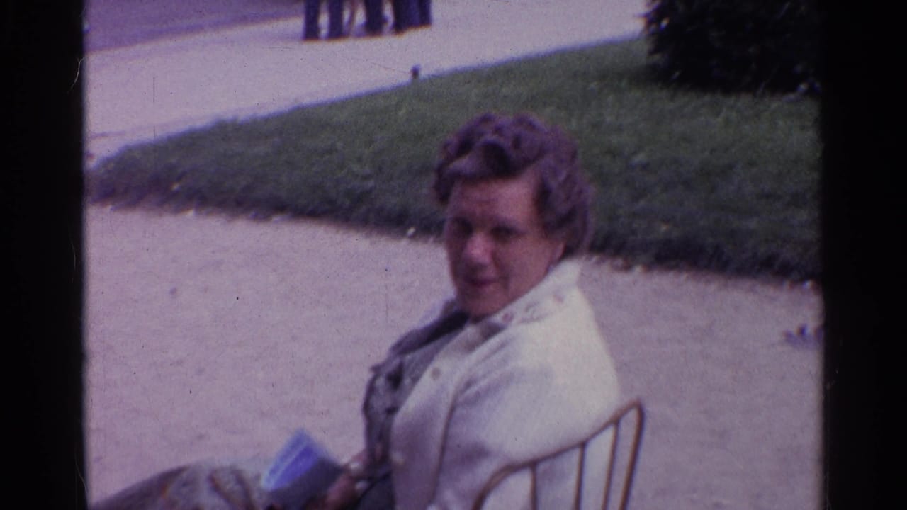 A woman in a light-colored jacket sits near a walkway and lawn in 1960s 8mm travel footage.