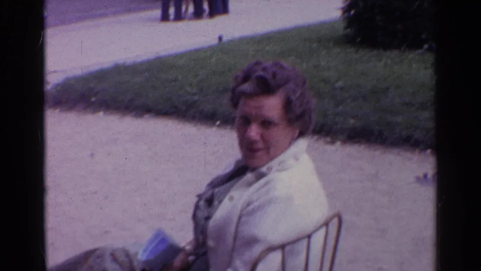 A woman in a light-colored jacket sits near a walkway and lawn in 1960s 8mm travel footage.