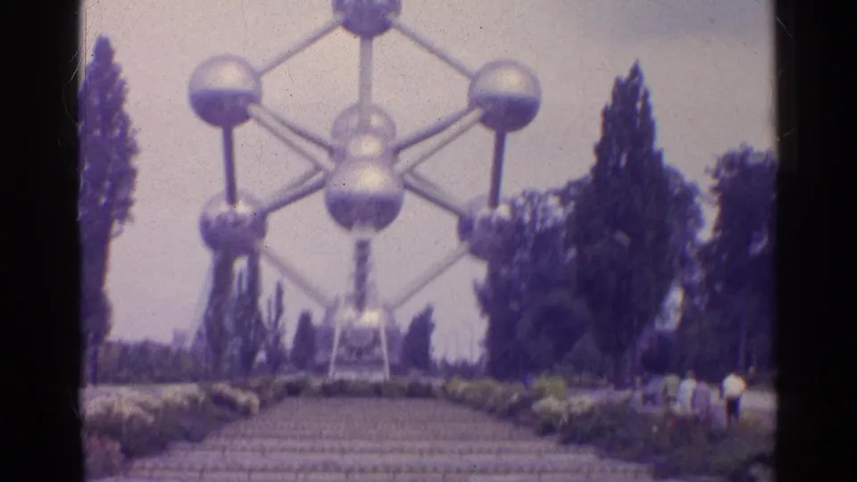 1961 8mm frame of a large silver interconnected-sphere landmark, with a path, trees, and distant visitors.