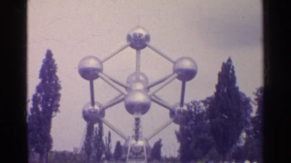 1961 8mm frame of the sphere-and-rod landmark centered between tall trees, with a small visitor near the lower edge.