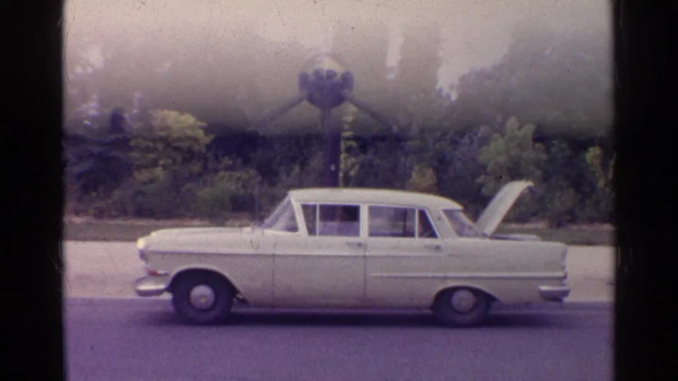 1961 8mm frame of a white classic sedan parked beside trees, with the trunk lid raised.