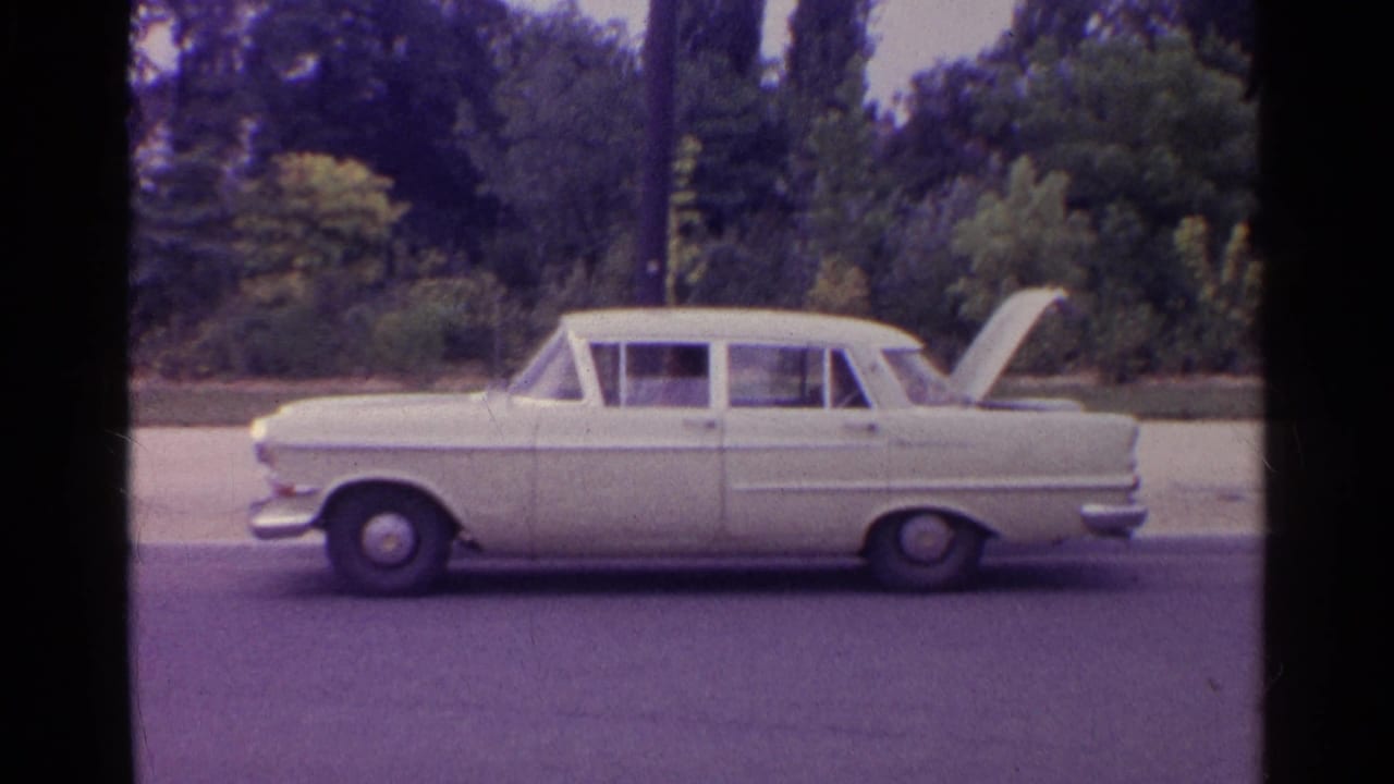 1961 travel footage frame showing a white sedan in profile with its trunk open near roadside greenery.
