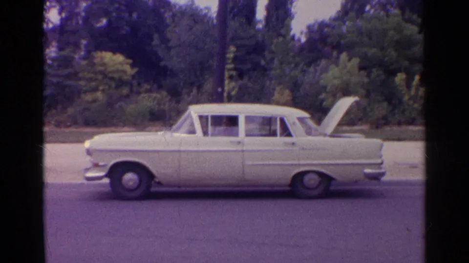 1961 travel footage frame showing a white sedan in profile with its trunk open near roadside greenery.