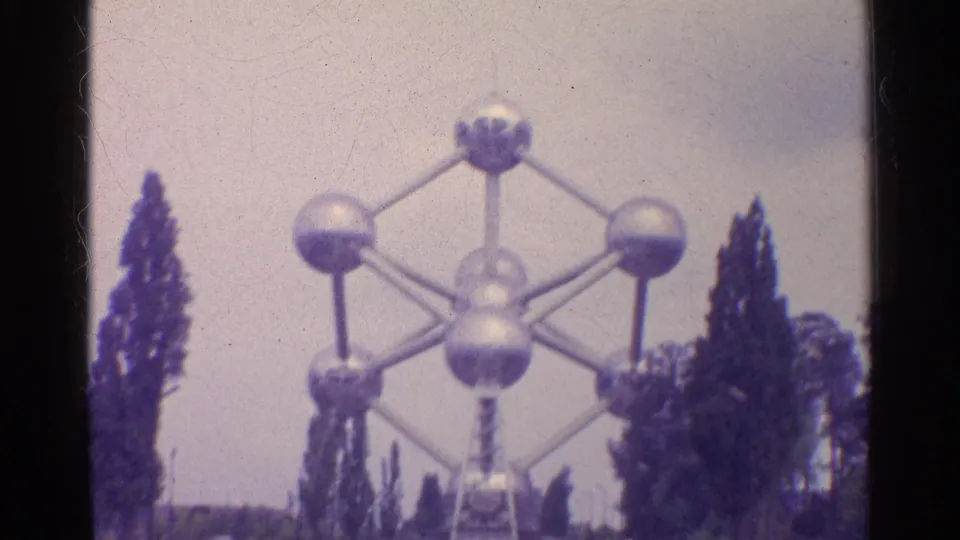 1961 8mm frame showing a large atom-like landmark structure rising above trees in a travel setting.