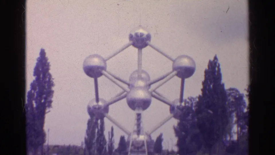 1961 home-movie frame of an atom-like metal landmark structure seen through trees, with faded color and soft focus.