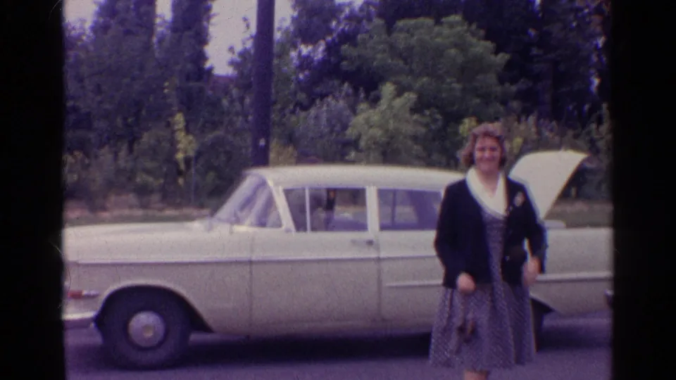 A woman in a patterned dress and dark cardigan stands by a light-colored 1960s sedan during a roadside stop.