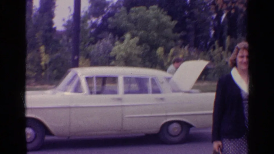 A light-colored sedan sits with its hood open while a woman passes at the edge of the 1961 8mm frame.