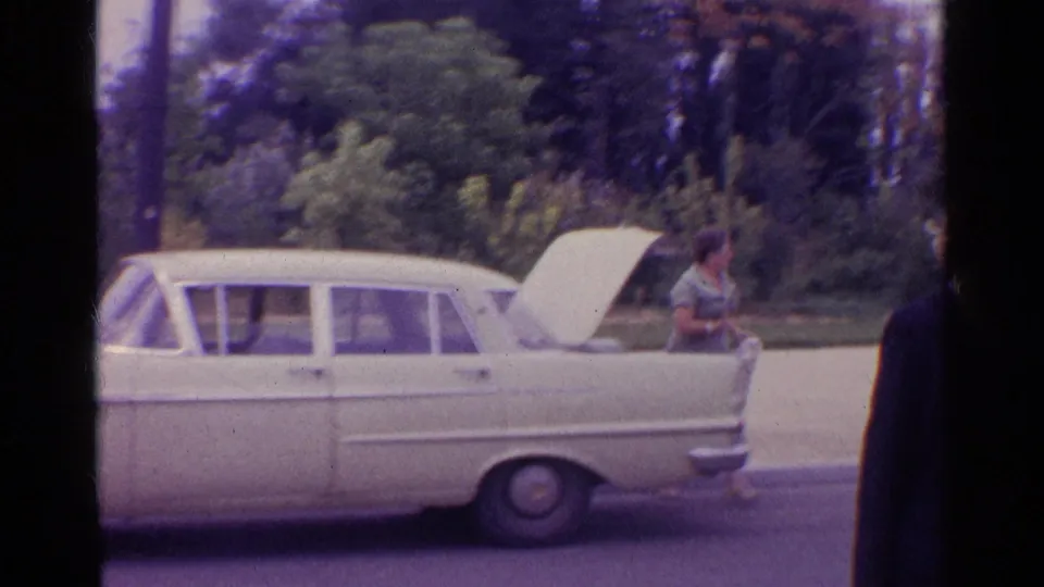 1961 home-movie frame showing a pale mid-century sedan with its hood raised beside trees and a person tending to the front of the car.