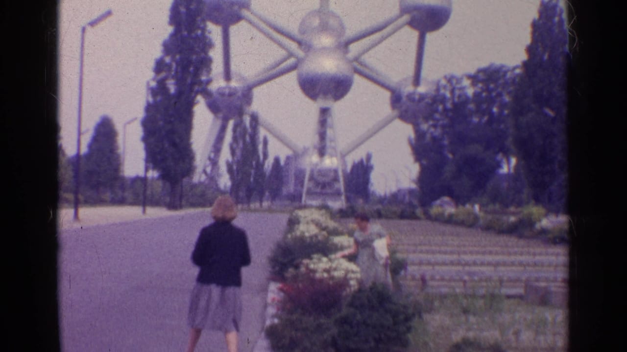 A woman in a grey skirt walks through landscaped grounds toward a large silver sphere-and-strut landmark, with another visitor near the steps.