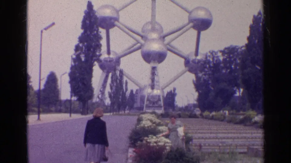The camera frames the monumental metal structure beyond a paved approach, with a woman walking away and another person near the landscaped steps.