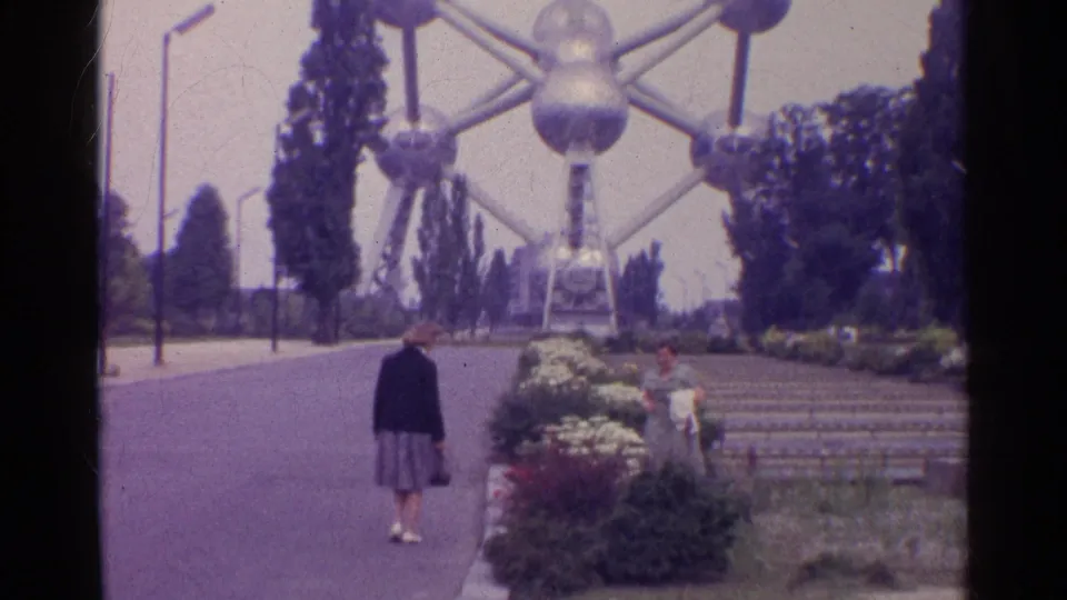 A woman in a dark jacket and grey skirt approaches landscaped steps beneath the large silver sphere-and-strut landmark, with another visitor at right.