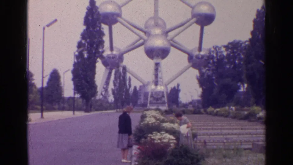 A woman stands on the paved approach while another person is near the flowerbeds below the Atomium in 1961 home-movie footage.