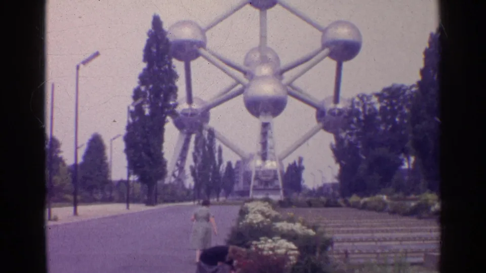 1961 8mm travel frame showing an Atomium-like landmark with trees, planted beds, paved paths, and a woman walking below.
