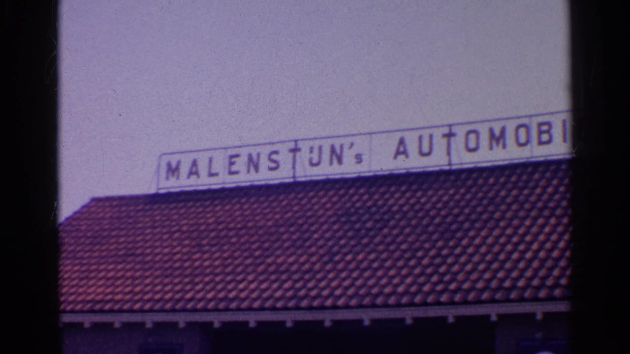 1961 8mm frame of a European automobile dealership sign above a red tiled roof, with dark film borders and soft focus.