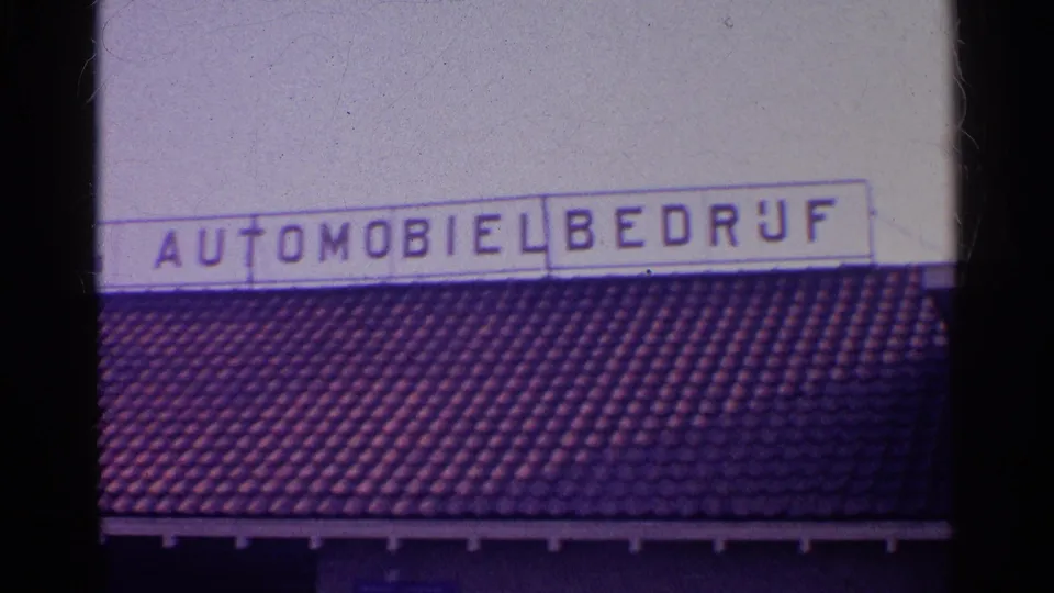 Tight 1961 8mm view of an automobile business sign over a tiled roof, with visible grain and slight exposure fade.