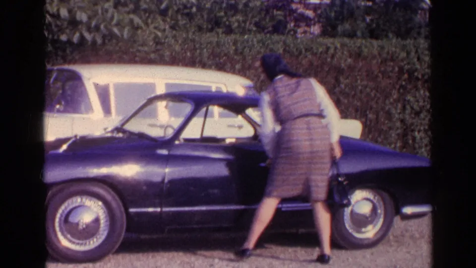 1961 Paris home-movie frame showing a woman beside a dark classic coupe, with a pale car and greenery in the background.