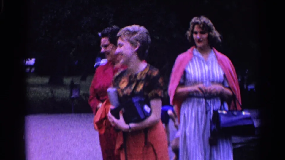 Three adult women in 1960s-style clothing stand together in a Swiss park, laughing and carrying handbags.