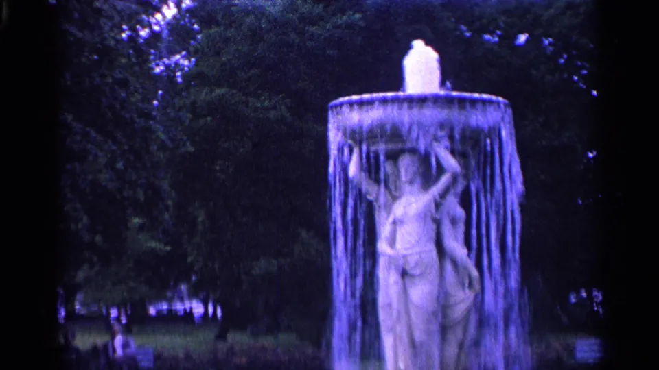 A classical fountain in a Swiss park, with water cascading around a pale sculpted figure and trees behind it.