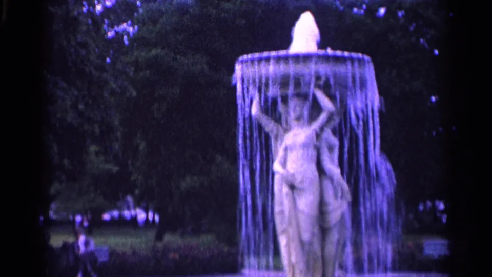 The fountain sculpture remains centered as water falls around it, with Swiss park greenery and benches visible behind.
