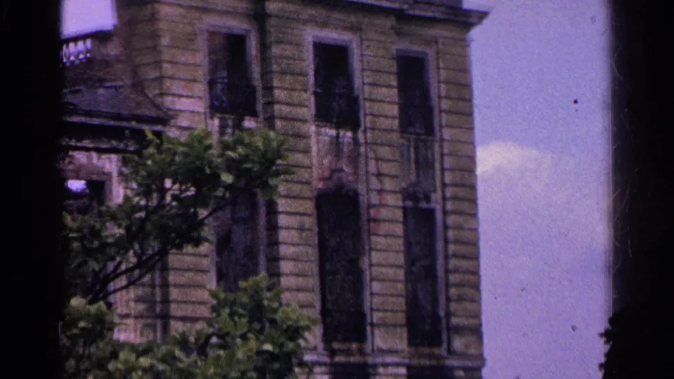 1961 Switzerland 8mm frame tilting lower on the historic building, showing tall windows, textured masonry, and tree branches.