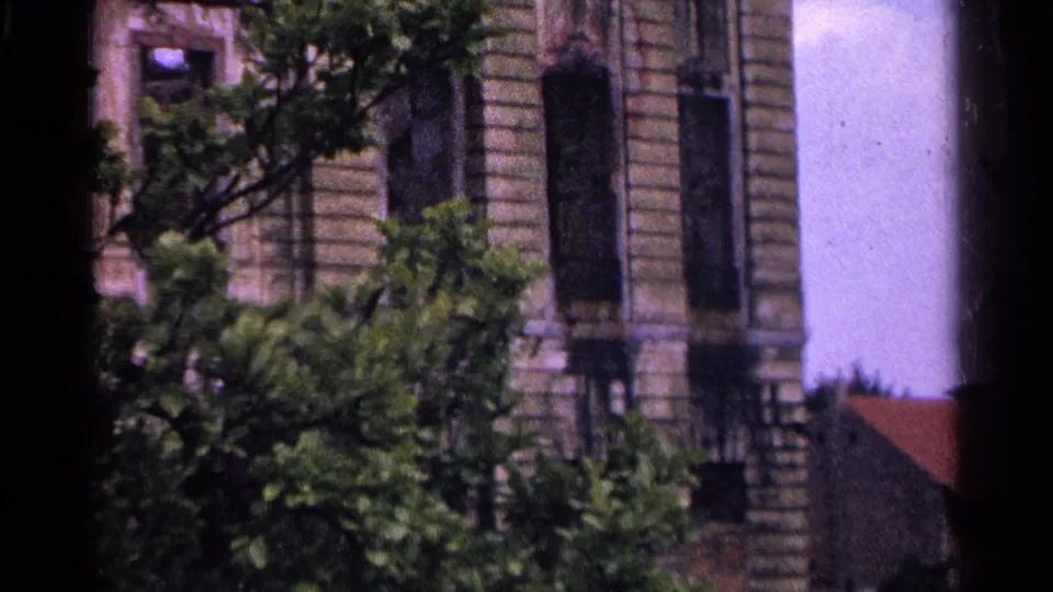 1961 Switzerland home-movie frame showing the lower facade of the ornate stone building, leafy trees, and a small red roof at the edge.