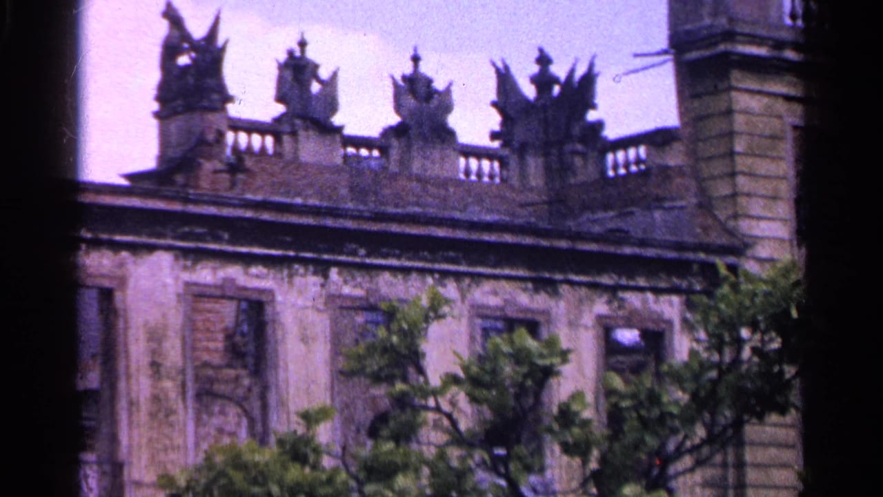 Roofline sculptures, shuttered windows, and a weathered facade seen above trees.