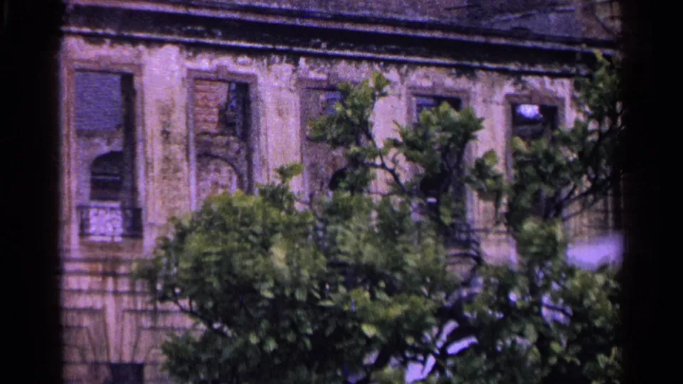 1961 Switzerland 8mm frame showing tall windows, closed shutters, and a weathered masonry facade partly blocked by trees.