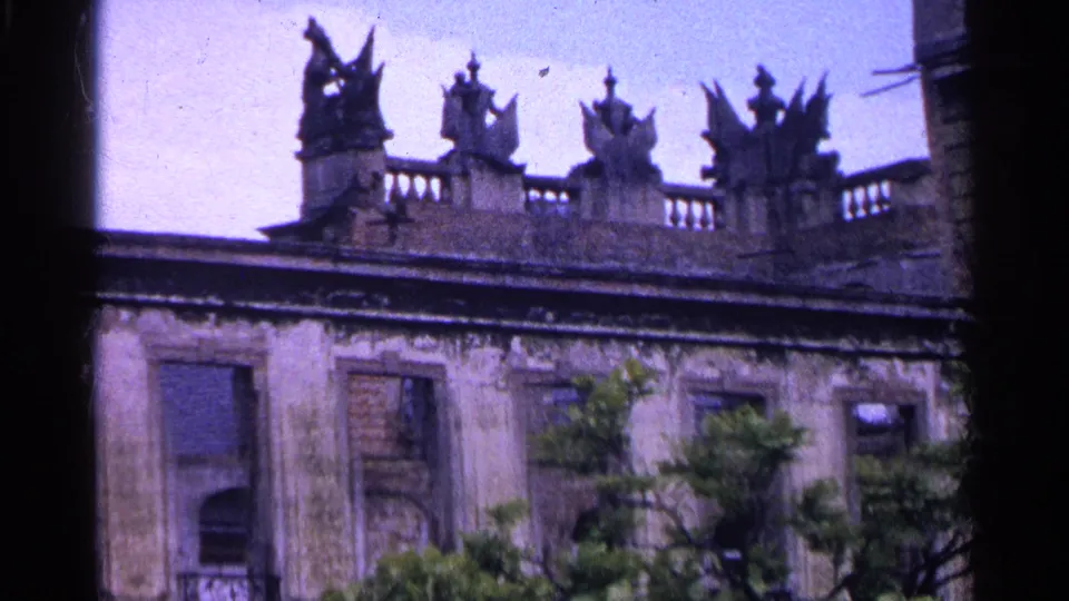 1961 Switzerland 8mm frame of a weathered ornate building roofline with statues, balustrade, and trees in the foreground.
