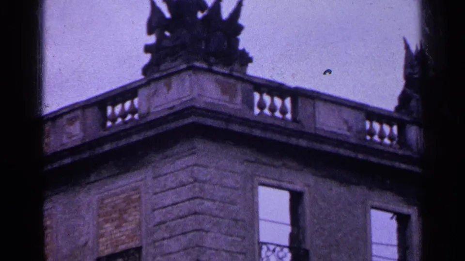 1961 Switzerland 8mm frame showing a stone corner, balustrade, open upper window, and dark roof statues against the sky.