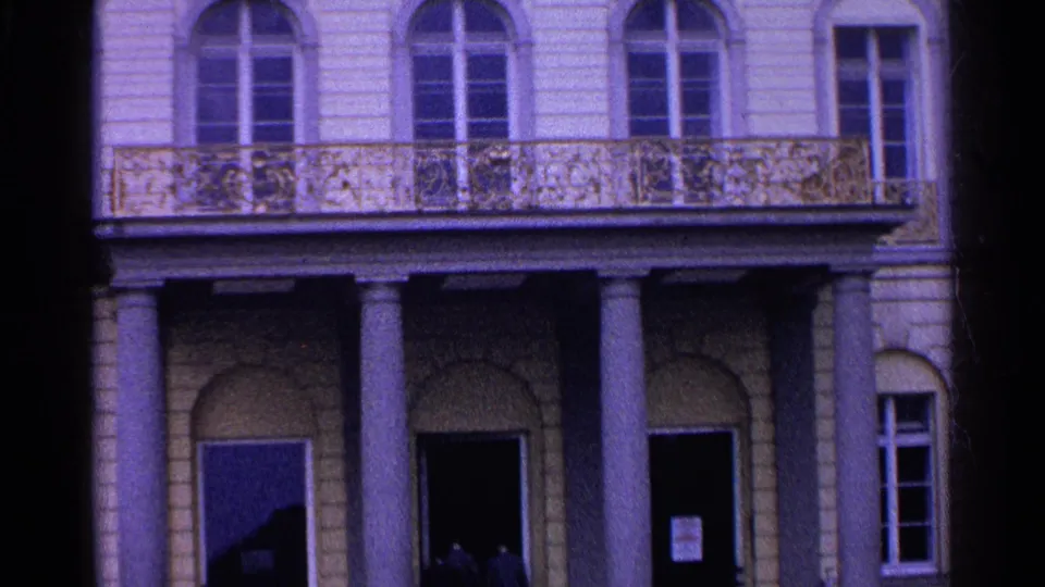 1961 Switzerland home-movie view of the lower facade, showing classical columns, a balcony, arched openings, and shadowed entrances.