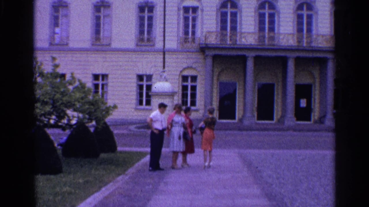 Four adults in 1961 Switzerland stand together on a paved courtyard before a pale classical building with arched doors.