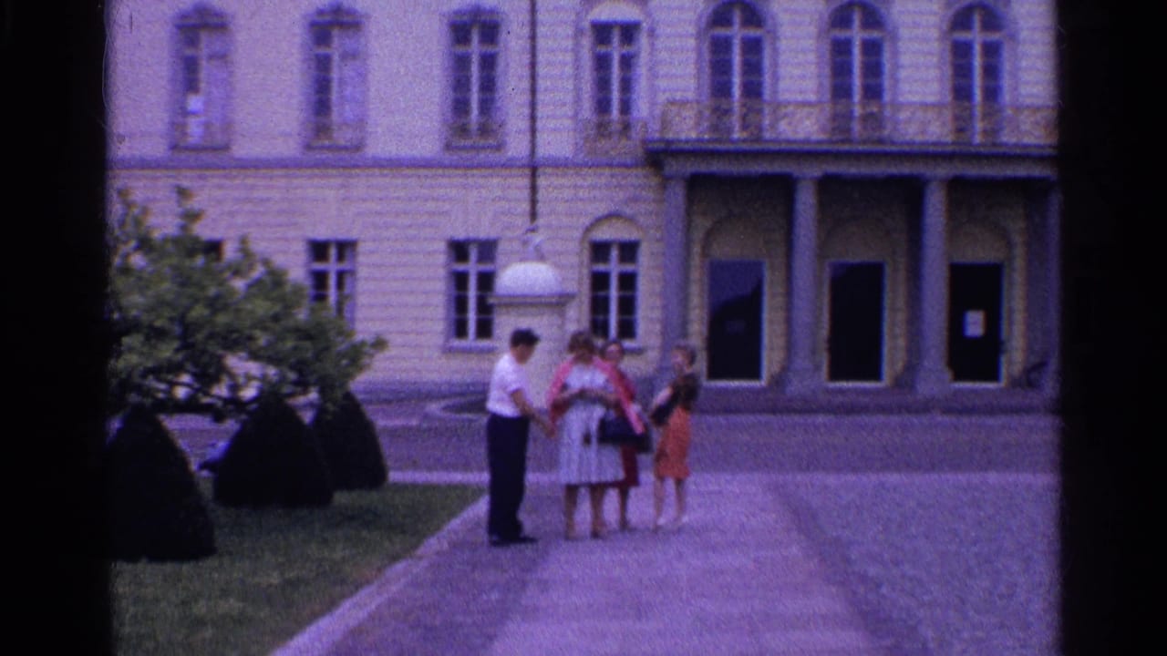 A man and three women appear to converse in a Swiss courtyard, with columns, arched doors, shrubs, and stone paving behind them.