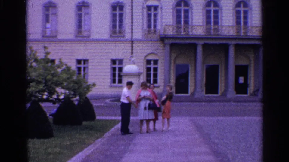 A man and three women appear to converse in a Swiss courtyard, with columns, arched doors, shrubs, and stone paving behind them.
