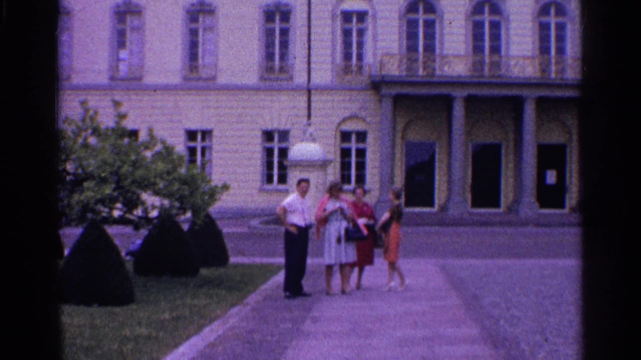 The 8mm frame shows four adults on a courtyard path in Switzerland, framed by shrubs and a pale classical building facade.