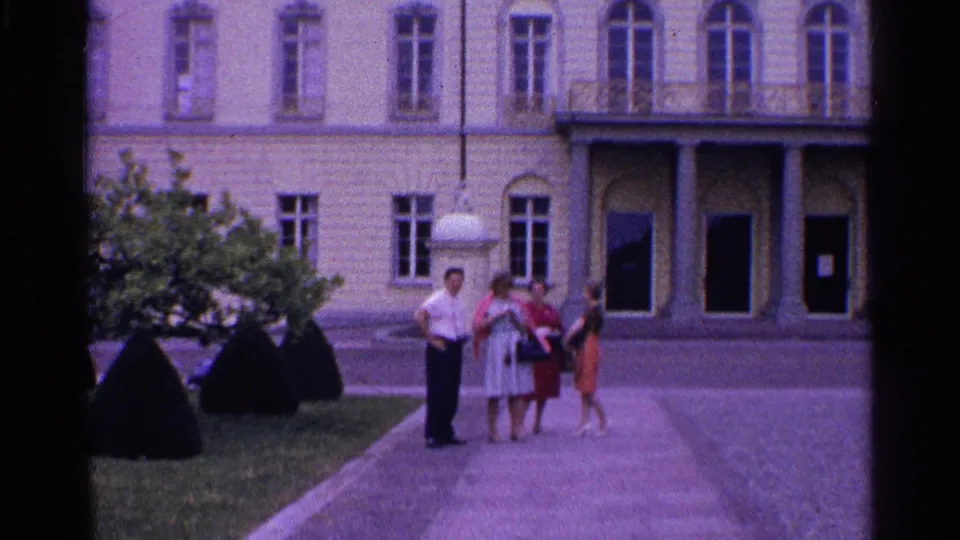 The 8mm frame shows four adults on a courtyard path in Switzerland, framed by shrubs and a pale classical building facade.