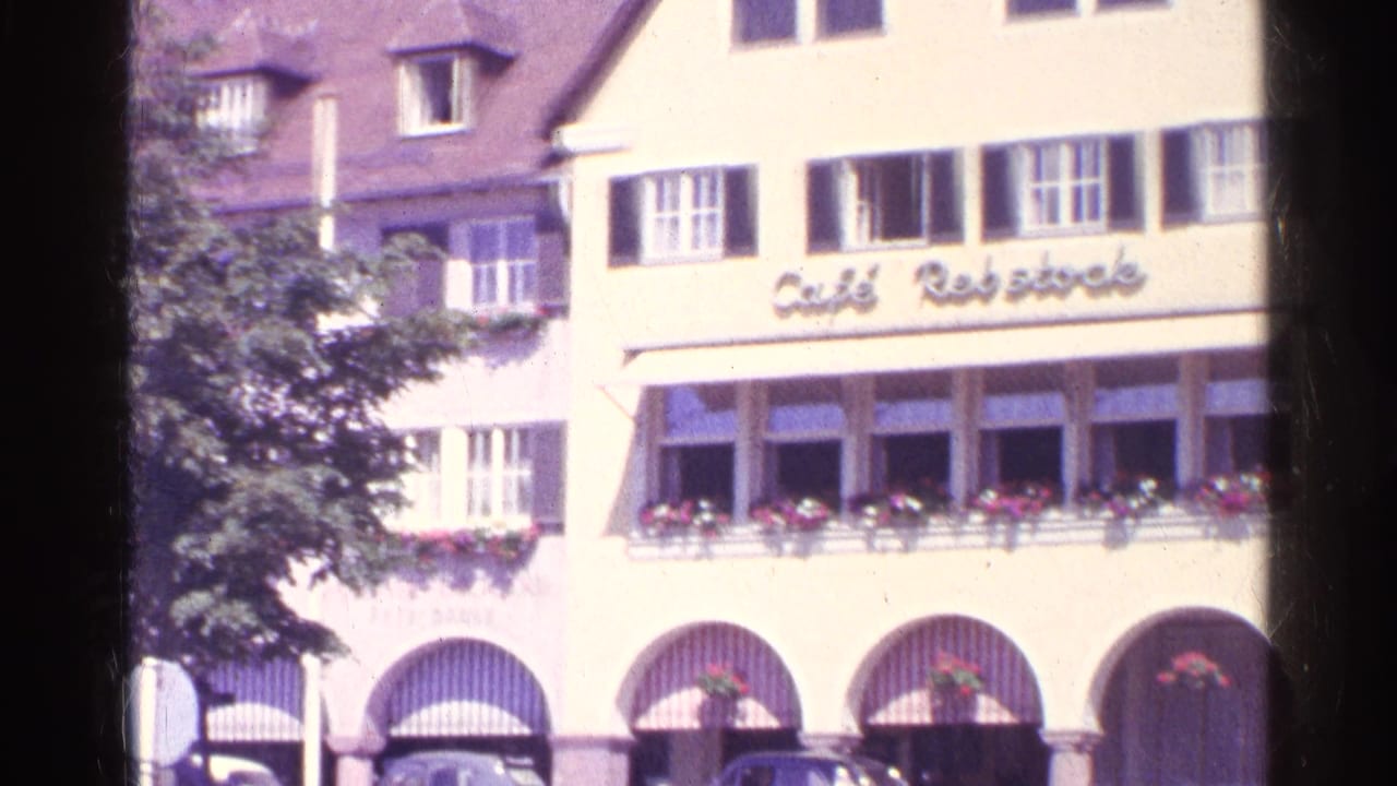 1961 Switzerland street frame showing a sunlit Cafe Rebsstock facade with upper shutters, flower boxes, and arched ground-floor openings.