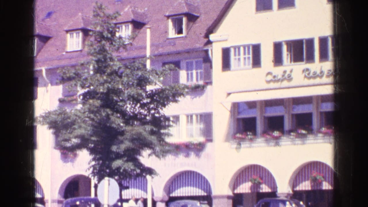 1961 Switzerland home-movie view of a shuttered cafe-side facade, with a leafy street tree partly covering windows and arched ground-floor bays.